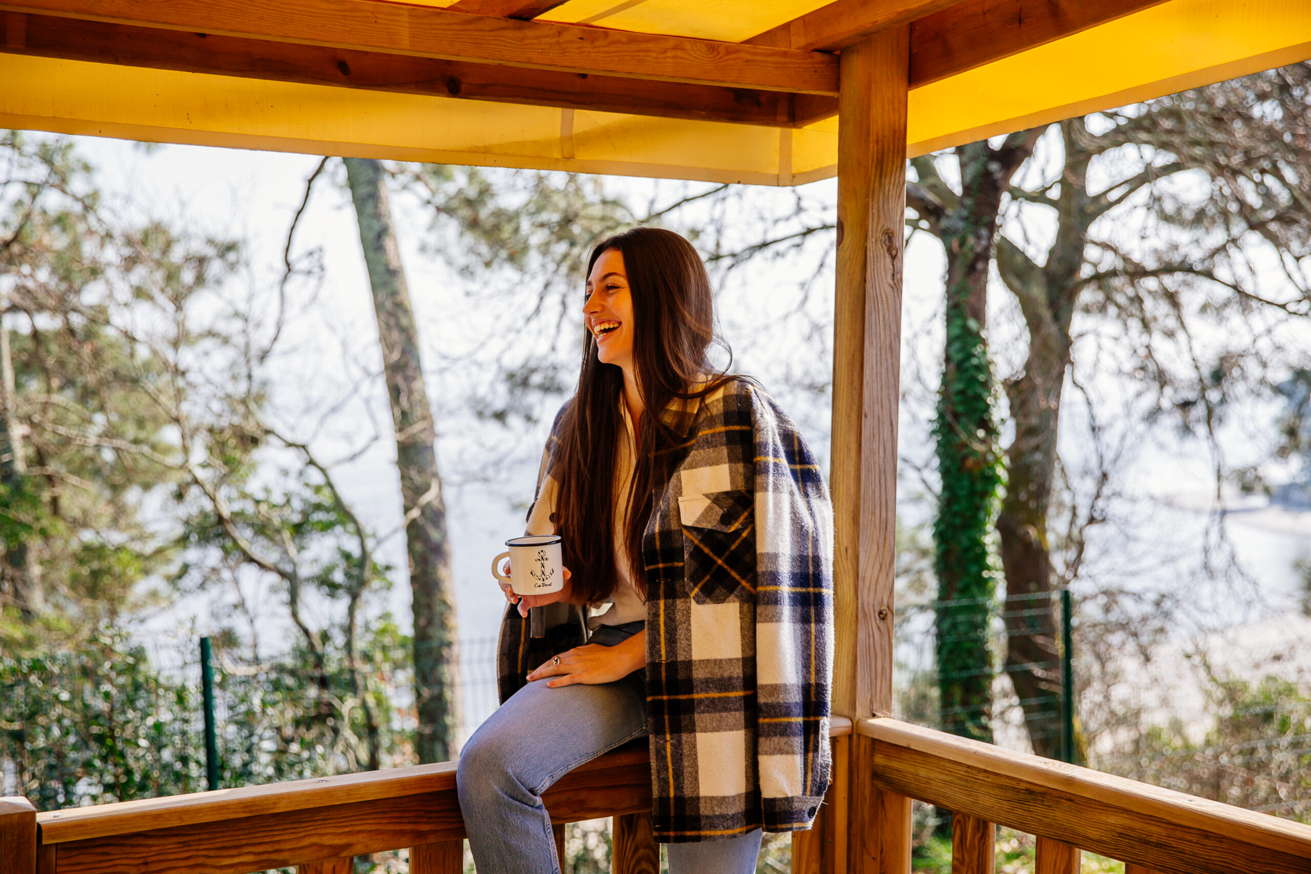 Une femme boit un thé assise sur la rambarde d'un mobil-home dans le camping les pastourelles, on peut également voir le bassin en arrière plan. 
