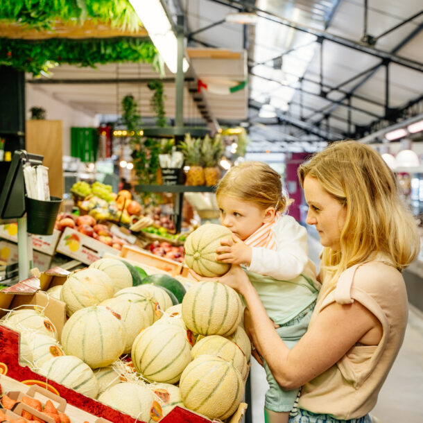 Une maman porte sa fille qui sent un melon sur un stand de fruits et légumes frais et locaux au marché couvert de Claouey à Lège-Cap Ferret, sur le Bassin d’Arcachon.