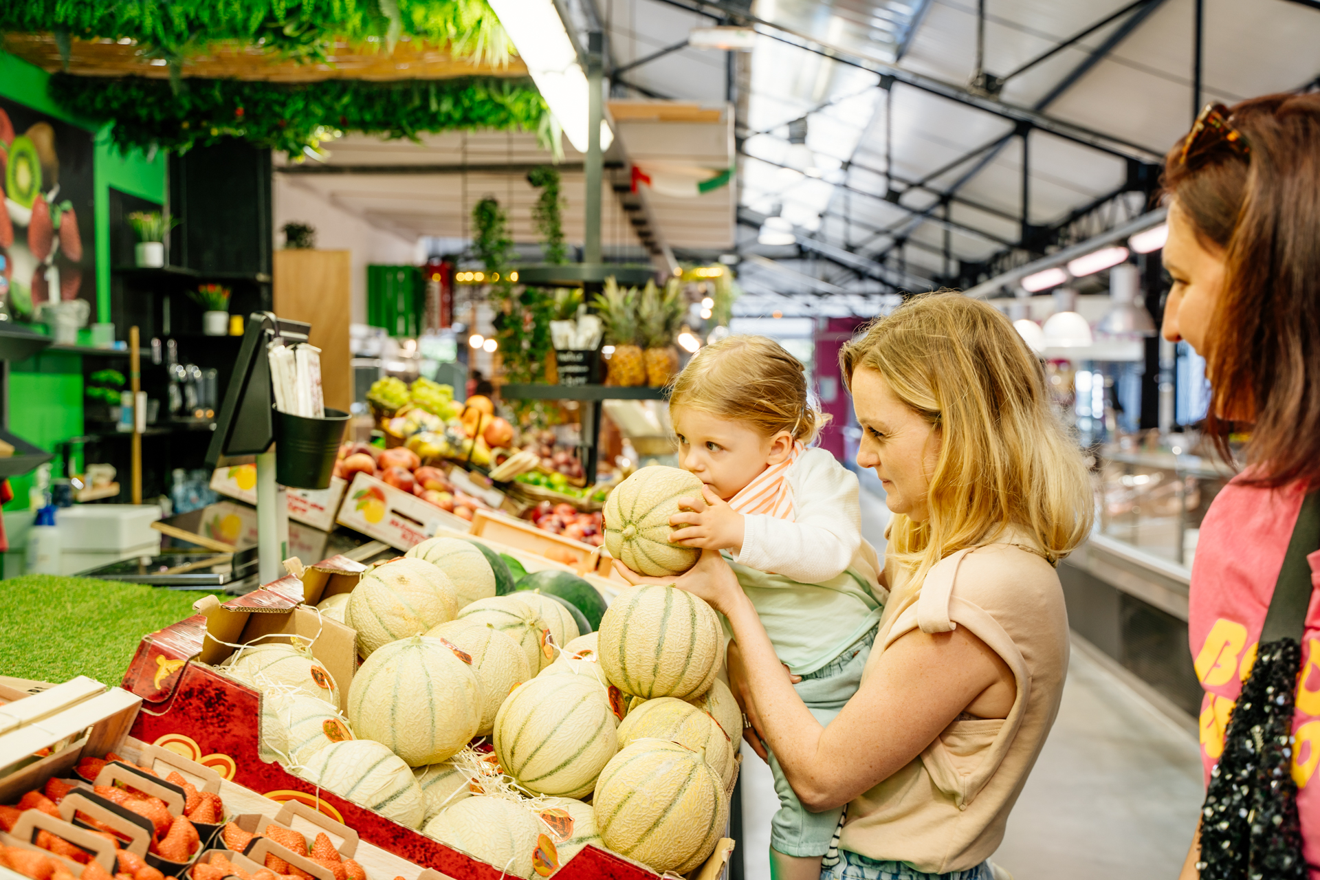 Une maman porte sa fille qui sent un melon sur un stand de fruits et légumes frais et locaux au marché couvert de Claouey à Lège-Cap Ferret, sur le Bassin d’Arcachon.