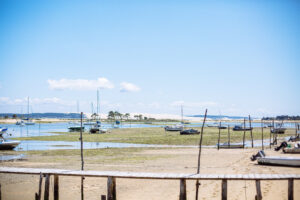 Vue sur une plage du bassin d’Arcachon à marée basse : des bateaux de plaisance reposent sur l’estran, des pieux et un ponton en bois apparaissent au premier plan, et une bande de sable bordée de pins se dessine à l’horizon sous un ciel bleu clair.