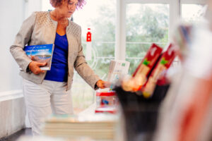 Une femme, livre à la main, parcourt la librairie du Phare du Cap Ferret. Vêtue d’une veste claire et d’un haut bleu, elle regarde des articles exposés sur une table au premier plan, tandis que des fenêtres laissent entrer la lumière en arrière‑plan. 