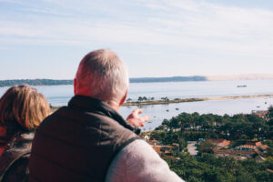  Deux personnes vues de dos observent le Bassin d’Arcachon depuis le sommet du Phare du Cap Ferret : l’homme, en gilet sombre, tend la main vers l’horizon où se dessinent des maisons, des pins, la lagune et une grande dune de sable au loin.